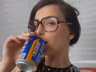 AlexandraCourtxxx holds a blue and orange energy drink can to her lips, showing her face and dark hair against a white blind background.