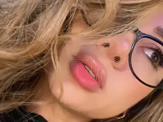 Alexavibe shows her face and blonde hair up close, looking into the camera with pink lips and dark-rimmed glasses.