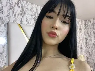 AleyahCoxx poses with long black hair and bangs, wearing a thin gold chain necklace against her bare skin.