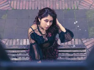 AliCipher poses with long dark hair, wearing a black lace top that shows her cleavage and shoulders while sitting on a brick bench.