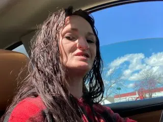 AliTaylorXXX has long, dark wavy hair and is looking at the camera while sitting in a car seat.