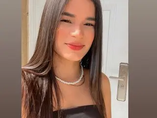 Ali_Cross20 poses with long dark hair and red lipstick, wearing a thin silver necklace and a black top.