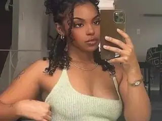 Alibabex22 poses for a mirror selfie, showing her face, dark curly hair, and a light green tank top.