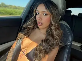 AliceHarriss poses with long wavy hair, wearing an orange top while looking directly at the camera in a car seat.