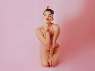 AliceKizar poses with her hands near her chest, wearing a light pink bikini against a solid pink background.