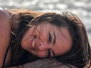 AliceRos smiles while lying down, showing her face and long brown hair against a soft, bright background.