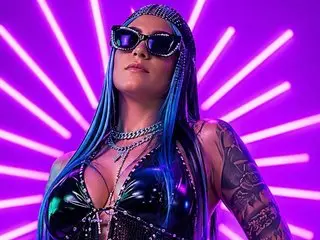 AliceSimpson poses with long blue hair, wearing a black leather bra and silver chain choker against a bright purple neon light background.