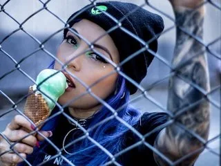 AliceSimpson with blue hair and a black beanie eats a mint ice cream cone behind a chain-link fence.