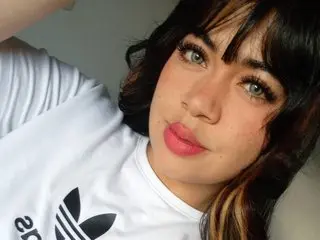 AliceTylor111 poses close-up, showing her face and long dark hair with bangs, wearing a white t-shirt with a black logo.
