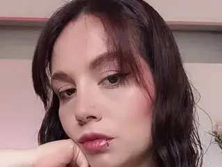 Aliison_Tyler poses close-up, showing her face and hair with a soft pink blush and dark wavy hair against a plain background.