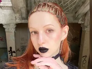 AlisaMoss with long red hair and black lipstick poses in front of a weathered building background.