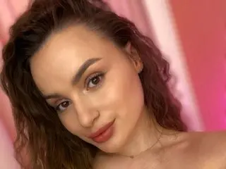 Young woman with wavy brown hair and glowing makeup posing in soft pink lighting.