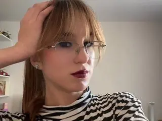 Young woman with blonde bangs and glasses wearing a striped turtleneck in a cozy indoor setting.