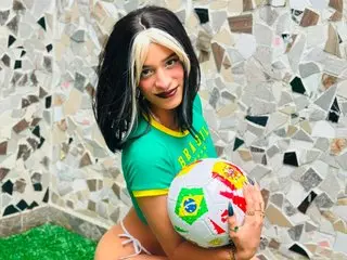 Alisha_Evans_Rs, with long black and blonde hair, poses in a green top while holding a soccer ball against a mosaic tile background.