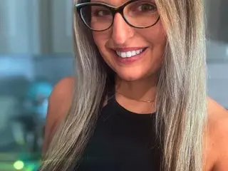 Smiling woman with long grey hair and glasses wearing a black tank top in a bright indoor setting.