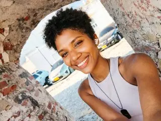 Smiling young woman in a grey tank top taking a selfie inside a rustic stone archway on a sunny day.
