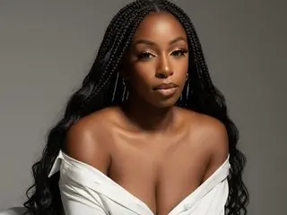 AliviaBlaze poses with her long black braids draped over her shoulders, wearing a white open-neck top against a plain grey background.