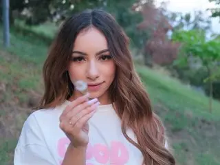 AlizeBlair poses with her long brown hair and a white t-shirt, looking directly at the camera against a soft, leafy outdoor background.