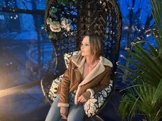 Alleyesonm poses looking away, wearing a brown jacket and light blue jeans, with her hair down in a dark outdoor setting.