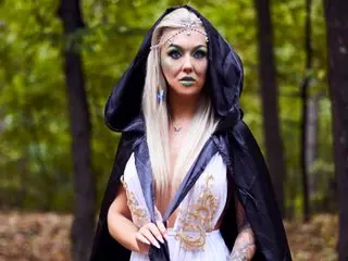 AllyRey poses in a black hooded cloak and white lace top, showing her long blonde hair and light makeup against a forest background.