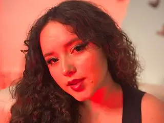 Young woman with curly hair and glamorous makeup posing in warm red lighting.