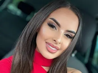 Smiling woman with long dark hair and glamorous makeup wearing a red top inside a car.