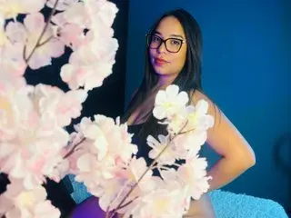 AlunaClark wearing glasses and a black top, posing behind white flowers against a blue background.