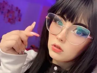 Alyssa_Romanova poses with a direct gaze, wearing large clear-rimmed glasses and a white top with her long black hair styled with blunt bangs.