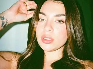 AmaiaSalvatore poses with her hand near her face, showing her long dark hair and clear skin against a plain background.