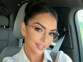 AmaliaRosse poses in a white collared shirt with her dark hair pulled back in a bun, looking directly at the camera while sitting in a car seat.