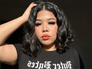 AmandaMoore1111 poses with her hands in her dark, wavy hair, wearing a black graphic t-shirt against a dark background.