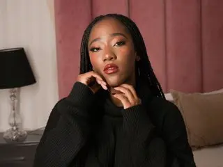 AmaraHofman poses with her hands near her neck, wearing a black ribbed sweater and showing her long dark braids against a pink background.