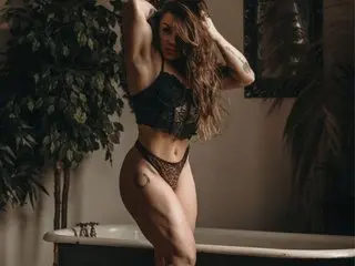 AmaraJezebel poses naked with long brown hair, showing her bare breasts and pussy while standing near a leafy plant and a white bathtub.