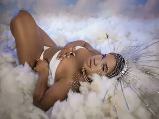AmaraSexxx lies back in a cloud-like setting, wearing a white bikini top and a large, decorative headpiece while showing off her tanned skin.