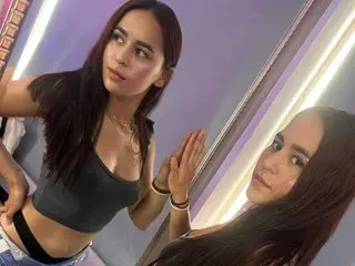 Amaratyche poses with long dark hair, wearing a dark grey tank top that shows her cleavage, looking directly at the camera.