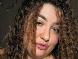 Young woman with long curly brown hair and natural makeup smiling in a cozy indoor setting.