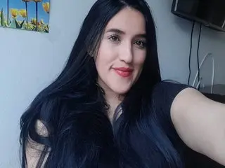 AmbarRossi poses with long dark hair and red lipstick, wearing a dark top against a plain wall background.