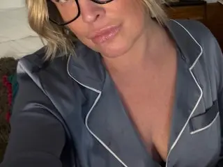 Blonde woman in grey satin pajamas with white trim relaxing in a cozy bedroom setting.