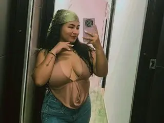 AmberMilers poses in a mirror wearing a tan bra and denim jeans, showing her cleavage and long dark hair.
