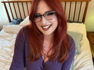 Smiling redhead woman in a purple top sitting on a bed in a cozy bedroom setting.