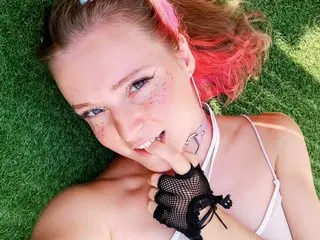 Smiling young woman with pink hair and freckles wearing a white top and black fishnet gloves on green grass.