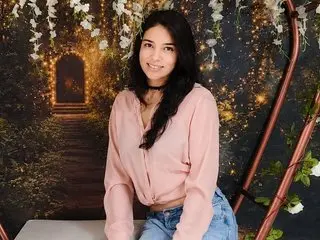AmelieSimon is a smiling woman with long dark hair wearing a pink long-sleeve shirt and blue jeans, sitting in front of a floral background.