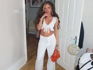 Amira_ayah poses in a white crop top and matching white trousers, showing her midriff and long wavy hair in a bright room.