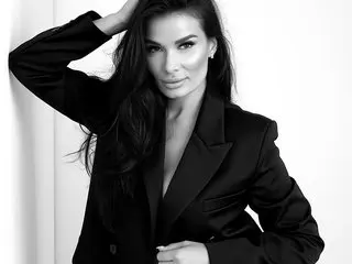 AmyChristie poses in a black blazer with her hands near her head, showing her long dark hair and bare chest in a black and white photo.
