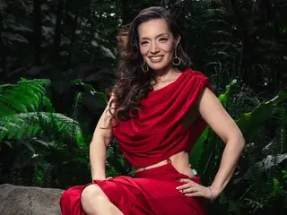 AmyRoxX poses with long wavy hair, wearing a red cut-out top and matching red skirt, set against a dark leafy jungle background.