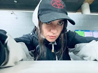AmySpider wearing a black and white baseball cap and a black zip-up jacket, looking directly at the camera with a serious expression.