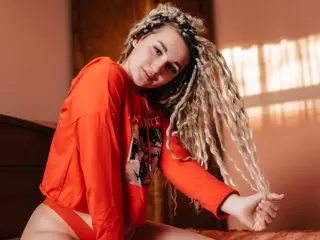 AmySwane with long blonde dreadlocks wears a red long-sleeve shirt and red panties while sitting on a bed.