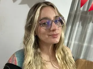 Smiling blonde woman with glasses and a gold necklace posing in a cozy indoor setting.