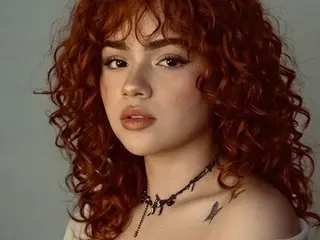 Anabel_Romero poses with her long, curly red hair and light makeup, showing her bare shoulders and neck with a black choker necklace.