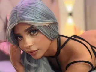 Anaiamoon111 poses with her long, light blue hair and dark eyes, wearing a black strappy top or bodysuit against a soft-focus background.
